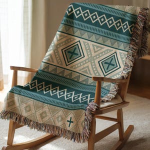May include: A woven throw blanket with a geometric pattern in shades of teal, beige, and white is draped over a wooden rocking chair. The blanket features diamond, triangle, and cross designs, with fringed edges. The chair is made of light-colored wood.