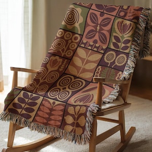 Può includere: Una coperta tessuta con un motivo floreale geometrico nei toni del marrone, viola e verde è drappeggiata su una sedia a dondolo in legno. La coperta ha un bordo con frange e un design patchwork con varie forme botaniche stilizzate.