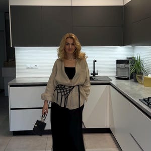 Może przedstawiać: Osoba w beżowej koszuli, czarnym topie i czarnej spódnicy stoi w nowoczesnej kuchni. Ma na sobie czarny pasek i małą czarną torebkę. Kuchnia posiada białe i ciemnoszare szafki.