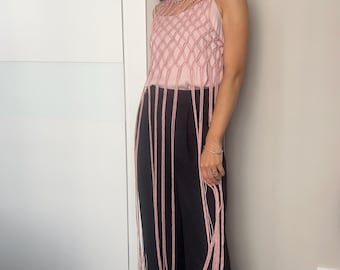 May include: A pink macrame top with fringe detail is paired with black pants. The top has a woven design on the upper portion and long fringe that hangs down. The outfit is completed with light pink sandals.