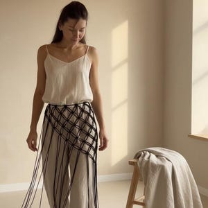 Pode incluir: Uma mulher usando uma camisola creme e calças de perna larga. Um cinto de macramê preto com franjas longas é usado sobre as calças. Um banco de madeira com um cobertor drapeado está ao fundo.