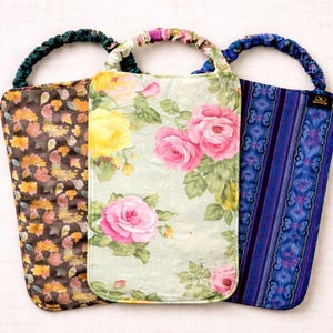 May include: Three fabric wine bottle bags with handles. The bags feature different floral and patterned designs. One bag has a brown floral pattern, another has pink and yellow roses, and the third has a blue and purple pattern.