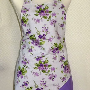 May include: A white apron with a purple floral print and a solid purple bottom panel. The apron is on a white mannequin.