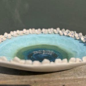 May include: A shallow ceramic bowl with a smooth, glossy interior featuring a gradient of deep blue at the centre transitioning to light turquoise. The outer rim is decorated with a continuous border of small, white, sculpted flower petals.