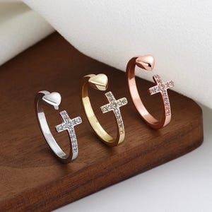 May include: Three open-ended rings with a cross design. One ring is silver, one is gold, and one is rose gold. Each ring features a heart-shaped end and a cross embellished with small, clear stones.