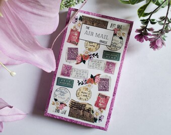 May include: A pink and white paper journal with a collage of vintage postage stamps featuring the words "The Air Mail Oracle" and "Pray for Peace".