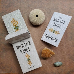 May include: The Wild Life Tarot deck and guidebook feature a white background with black and gold text and illustrations. The deck box has a white background with black and gold illustrations of a snake and a star. The guidebook has a white background with black and gold illustrations of a snake and a star.
