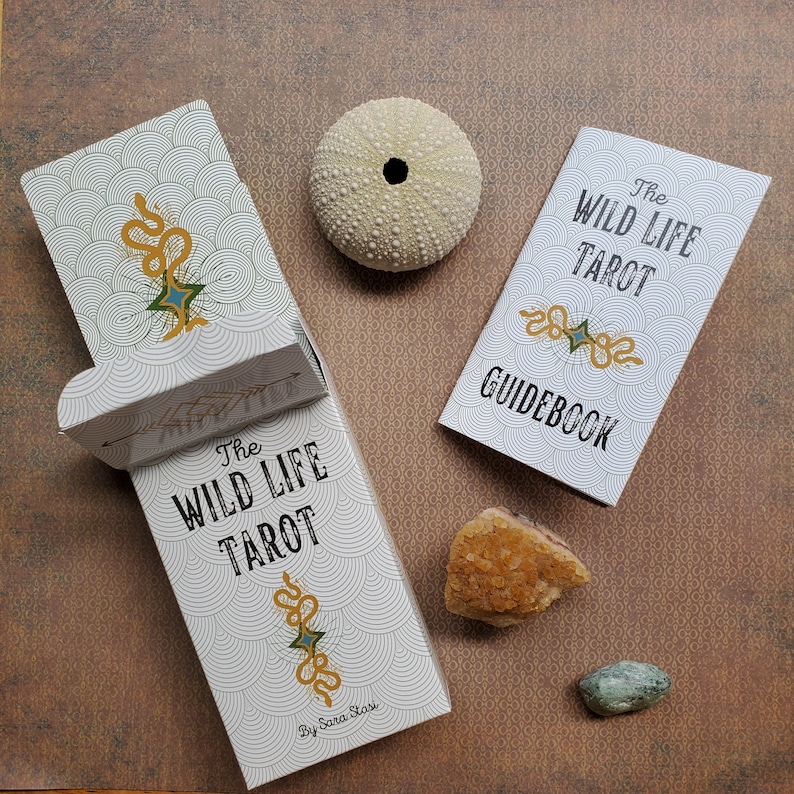 May include: The Wild Life Tarot deck and guidebook feature a white background with black and gold text and illustrations. The deck box has a white background with black and gold illustrations of a snake and a star. The guidebook has a white background with black and gold illustrations of a snake and a star.