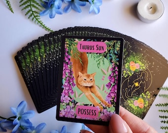 May include: A deck of tarot cards featuring a card with a stretching orange cat illustration. The card reads "Taurus Sun" and "Possess" with floral accents. The deck is black with a green and gold design. Blue flowers and a candle are in the background.