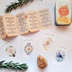 May include: A set of tarot cards with floral designs and the words "The Hearth Home Tarot" on the tin box. The cards feature images of brooms, pentacles, wands, and cups.