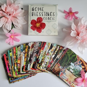May include: A deck of oracle cards fanned out, featuring colorful photographic imagery, alongside a rectangular box labeled HOME BLESSINGS ORACLE with a red flower illustration, surrounded by several light pink artificial flower blossoms.