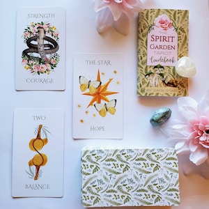 May include: A set of tarot cards with illustrations of a snake, a star, and a branch. The cards are white with black text and colorful illustrations. The cards are laid out on a white surface with a book titled "The Spirit Garden Tarot Guidebook" by Sara Stasi.