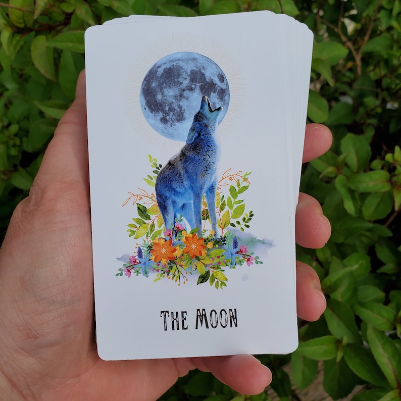 May include: A tarot card with a blue wolf howling at a full moon. The card is surrounded by flowers and greenery. The text "THE MOON" is printed at the bottom of the card.