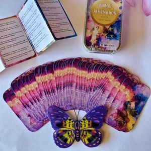 May include: A set of 42 animal affirmation oracle cards in a purple and pink watercolor design. The cards are fanned out in a semi-circle with a purple butterfly in the center. The cards are in a tin box with the text "Animal Affirmation Oracle Card Deck" and "By Sara Stasi".