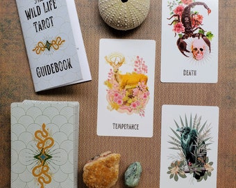 May include: A set of tarot cards with a guidebook, featuring a variety of illustrations. The cards include images of a scorpion, a deer, a bird, and a tower. The guidebook has the title "The Wild Life Tarot Guidebook".