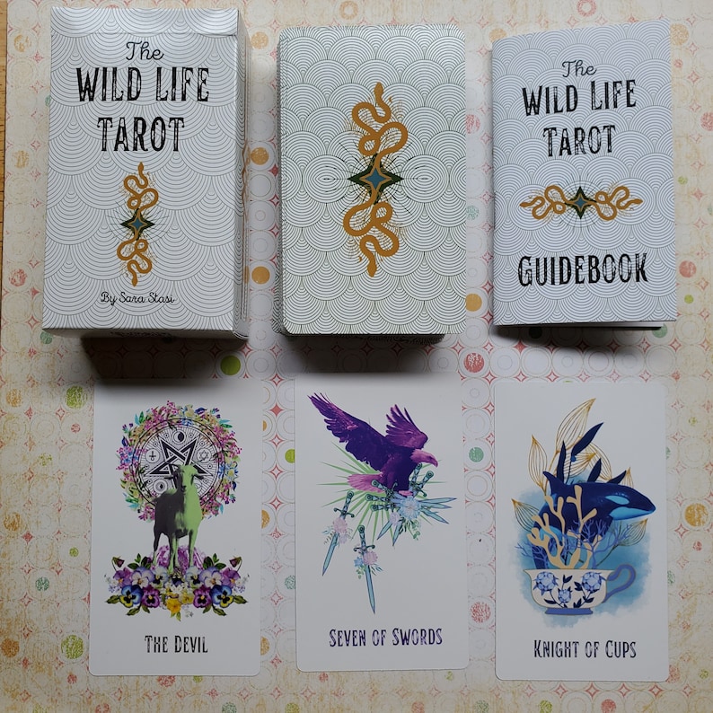 May include: The Wild Life Tarot deck and guidebook by Sara Stasi. The deck features colorful illustrations of animals and plants. The guidebook is white with green and gold accents.