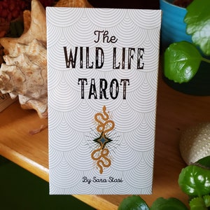May include: The Wild Life Tarot deck box features a white background with a repeating pattern of gray and white circles. The box has black text that reads "The Wild Life Tarot" and "By Sara Stasi". A gold snake with a blue star in the center is featured in the middle of the box.