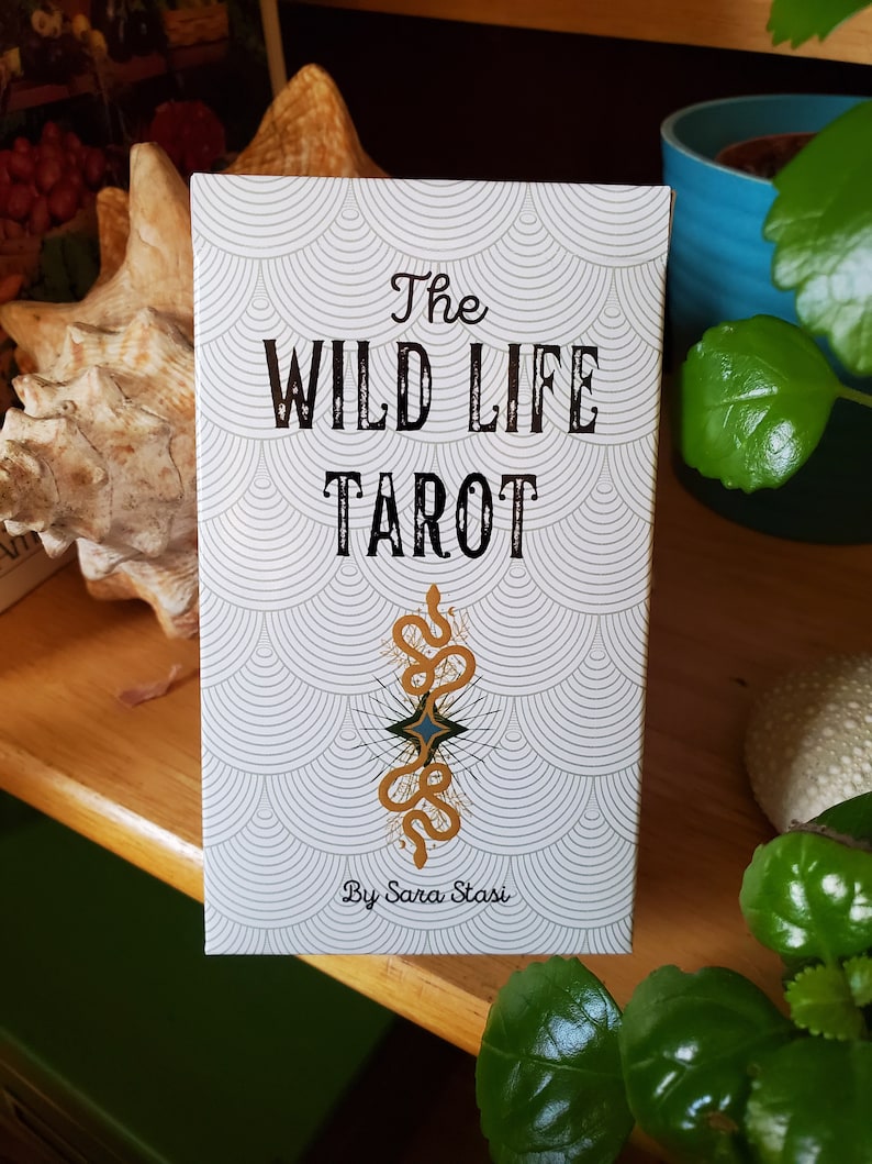 May include: The Wild Life Tarot deck box features a white background with a repeating pattern of gray and white circles. The box has black text that reads "The Wild Life Tarot" and "By Sara Stasi". A gold snake with a blue star in the center is featured in the middle of the box.