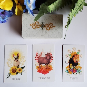 May include: Three tarot cards with colorful illustrations and gold accents. The Fool card features a white dog with a sunflower crown. The Emperor card features a ram with a crown. The Strength card features a lion with a crown.