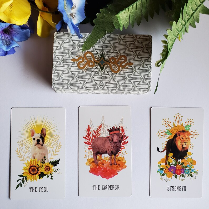 May include: Three tarot cards with colorful illustrations and gold accents. The Fool card features a white dog with a sunflower crown. The Emperor card features a ram with a crown. The Strength card features a lion with a crown.