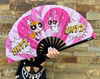 May include: Pink and white hand fan with a black handle. The fan features a cartoon image of a character from the Powerpuff Girls and a character from Hello Kitty. The fan has the word "UNTZ" printed on it.