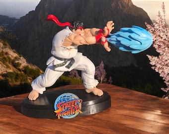 May include: A figurine of Ryu from Street Fighter, a popular video game series, in a fighting pose with a blue energy blast coming from his hand. The figurine is on a black base with the Street Fighter logo.