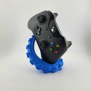 Può includere: Un controller Xbox nero è tenuto da un supporto blu a forma di tentacolo. Il controller ha pulsanti colorati e due joystick. Il supporto è di un blu vibrante e ha una forma ad anello con piccole protuberanze arrotondate.