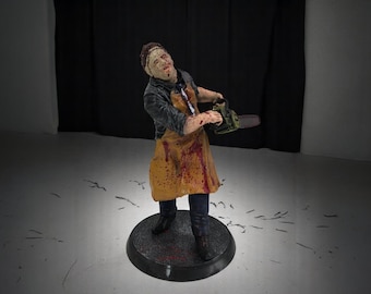 May include: A figurine of a person wearing a yellow and brown apron covered in blood splatters, holding a chainsaw. The figurine is standing on a black circular base.