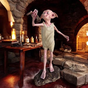 May include: A light-skinned elf figurine with pointed ears, wearing a white jumpsuit, stands on a rock with a wooden base. The elf is holding a wooden stick in its right hand and is looking to the right. The figurine is in a dark, stone room with a fireplace in the background.