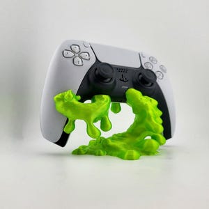 May include: A white and black PlayStation 5 controller with a neon green, dripping slime design. The controller has a black touchpad and buttons, with the PlayStation logo visible. The slime appears to be flowing from the bottom of the controller.