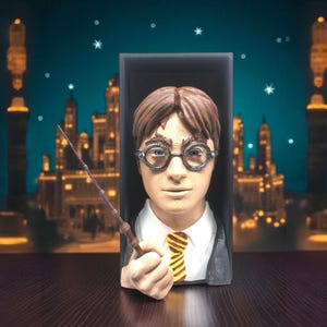 Könnte beinhalten: Detaillierte Büste einer Figur mit braunem Haar, runder Brille und einer Blitznarbe. Die Figur trägt ein weißes Hemd, eine gelb-schwarz gestreifte Krawatte und eine dunkle Jacke. Die Büste hält einen Zauberstab. Der Hintergrund zeigt ein Schloss.