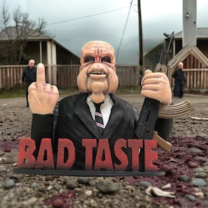 May include: A sculpture of a figure in a suit and tie, holding a rifle and giving the middle finger. The word "BAD TASTE" is displayed in red block letters at the base of the sculpture. The background shows a rural setting.