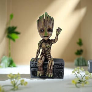 May include: A green and brown figurine of a character from the Guardians of the Galaxy movie series, sitting on top of a vintage boombox. The character is waving with one hand and has a friendly expression.