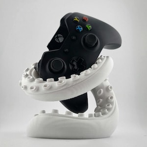 Peut inclure: Une manette Xbox noire est posée sur un support blanc en forme de tentacule. La manette comporte des boutons colorés et un pavé directionnel. Le support est conçu pour maintenir la manette en position verticale, ajoutant une esthétique unique.