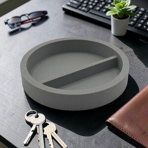 Peut inclure: Un organiseur de bureau gris de forme circulaire, divisé en deux compartiments. Fabriqué dans un matériau solide, il présente un design minimaliste. Des clés et un portefeuille sont visibles à proximité.