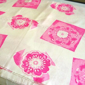 May include: White fabric with a repeating pattern of pink floral designs within square frames. The flowers have a detailed, layered petal structure and a lighter pink center. The fabric is folded, showing the texture and pattern.