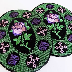 May include: Two oval-shaped black velvet patches with green and purple embroidery. Each patch features a pink and purple rose with green leaves and stems. The background of the patches is a green and black geometric pattern.