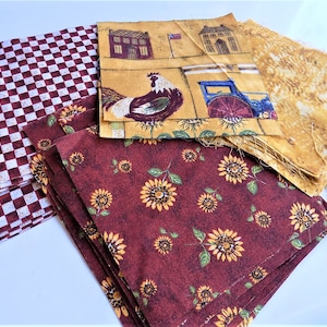 May include: A stack of fabric squares in various patterns, including red and white checkered, brown with yellow sunflowers, and yellow with a rooster and a house. The fabric squares are perfect for quilting or crafting projects.