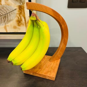 May include: A wooden banana holder with a curved design. The holder is made of natural wood and has a light brown finish. Four yellow bananas are hanging from the holder.
