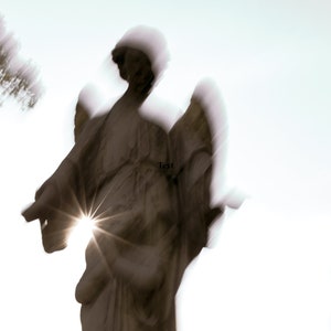 May include: A blurry image of a stone angel statue with wings, standing against a white background. The statue is illuminated by a bright light, creating a starburst effect. The text "Text" is visible on the statue.