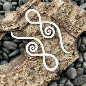 May include: Two white spiral hair clips with a simple design. The clips are on a piece of driftwood with gray stones in the background.