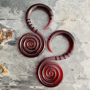 Könnte beinhalten: Zwei dunkelrote Ohr-Spiral-Dehner mit Farbverlauf und schwarzen Streifen. Die Dehner haben eine glatte, glänzende Oberfläche und ein Spiraldesign. Die Ohr-Dehner werden auf einer strukturierten, grauen Oberfläche präsentiert.