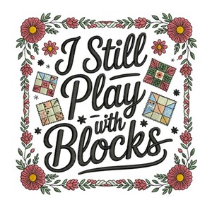 May include: Embroidered design with the text "I Still Play with Blocks" in black script, surrounded by a floral border with pink and yellow flowers and small quilt block illustrations. The design is on a white background.