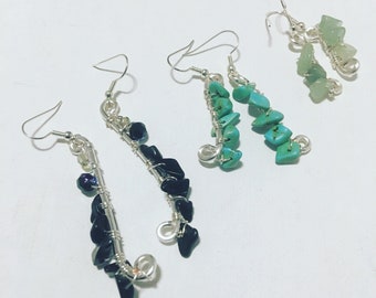 Puede incluir: Cuatro pares de pendientes hechos a mano con alambre de plata y detalles de piedras. Los pendientes presentan piedras negras, turquesas y verdes. Los pendientes están sobre una superficie blanca.