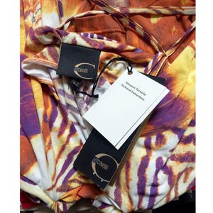 Op de afbeelding: Een Just Cavalli kledingstuk met een levendig abstract dierenpatroon in oranje, paars en wit. Het item bevat een zwart merklabel met een goudkleurig logo en een wit label met de tekst Istruzioni Tecniche Technical Instructions.