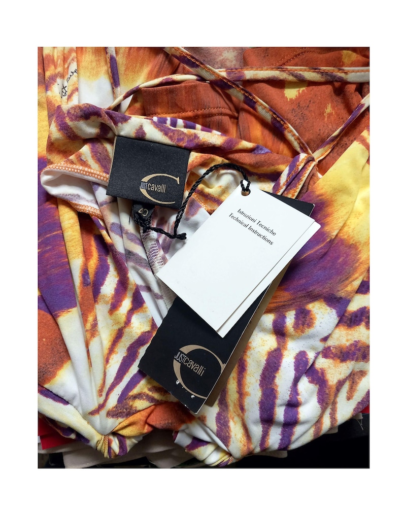 Op de afbeelding: Een Just Cavalli kledingstuk met een levendig abstract dierenpatroon in oranje, paars en wit. Het item bevat een zwart merklabel met een goudkleurig logo en een wit label met de tekst Istruzioni Tecniche Technical Instructions.