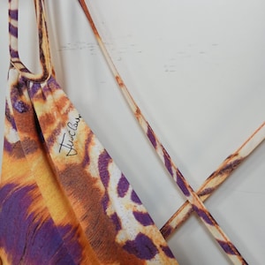 Op de afbeelding: Een mouwloos kledingstuk met een levendige dierenprint in tinten oranje, geel en paars. De stof is voorzien van dunne, kruisende spaghettibandjes en de tekst Just Cavalli in zwarte schrijfletters op het bovenste voorste gedeelte.