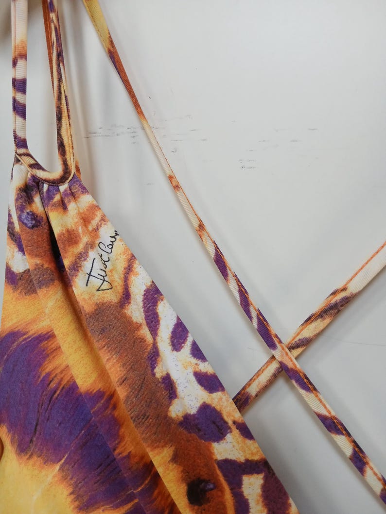 Op de afbeelding: Een mouwloos kledingstuk met een levendige dierenprint in tinten oranje, geel en paars. De stof is voorzien van dunne, kruisende spaghettibandjes en de tekst Just Cavalli in zwarte schrijfletters op het bovenste voorste gedeelte.