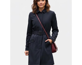 Op de afbeelding: Een marineblauwe trenchcoat met bijpassende riem en een bordeauxrode crossbodytas. De jas heeft een klassiek ontwerp met een knoopsluiting, zakken en een kraag. De riem is vastgemaakt met een rechthoekige gesp.