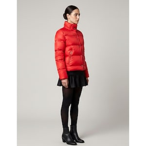 Puede incluir: Chaqueta acolchada roja brillante con cuello alto y botones dorados. La chaqueta se combina con una falda negra, medias negras y botines negros. La modelo está de pie frente a un fondo gris claro.